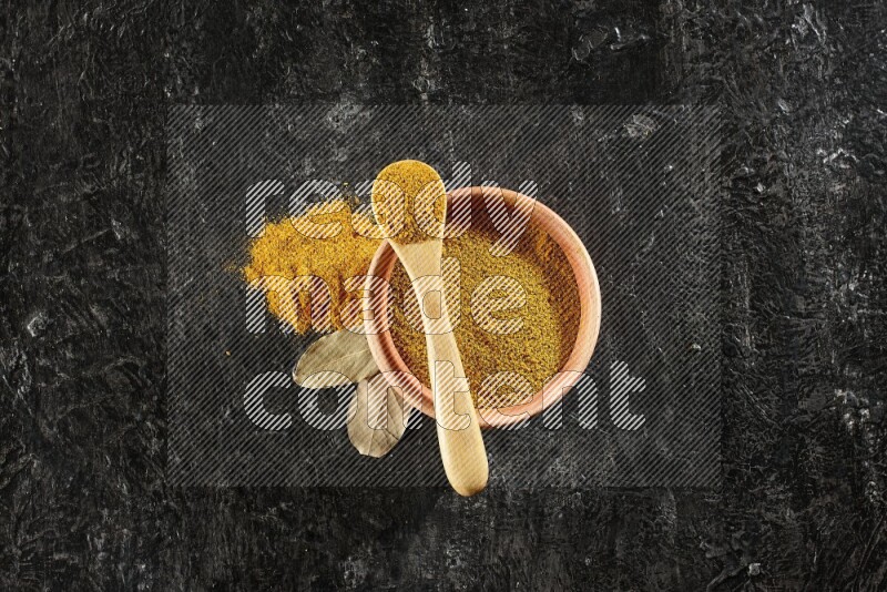 A wooden bowl and a wooden spoon full of turmeric powder on textured black flooring