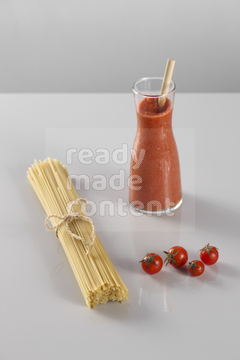 Raw pasta with tomatoe pasta with different ingredients such as cherry tomatoes, basil, garlic, bay laurel, cardamom, white pepper, black pepper, red chilis and wheat stalks on light grey background