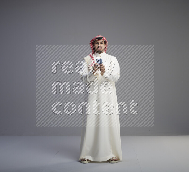 A  Saudi man standing wearing thob and red shomag texting on phone on gray background