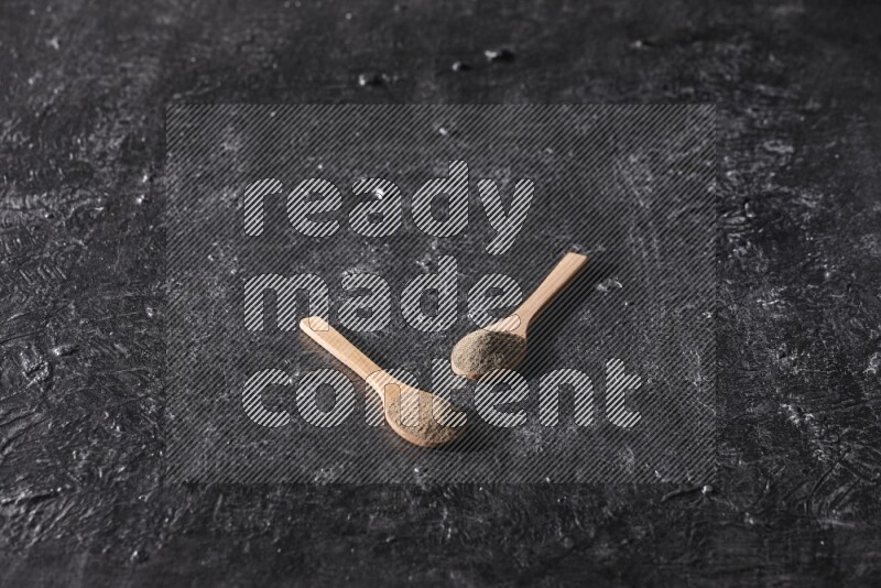 2 wooden spoons full of black pepper powder on a textured black flooring