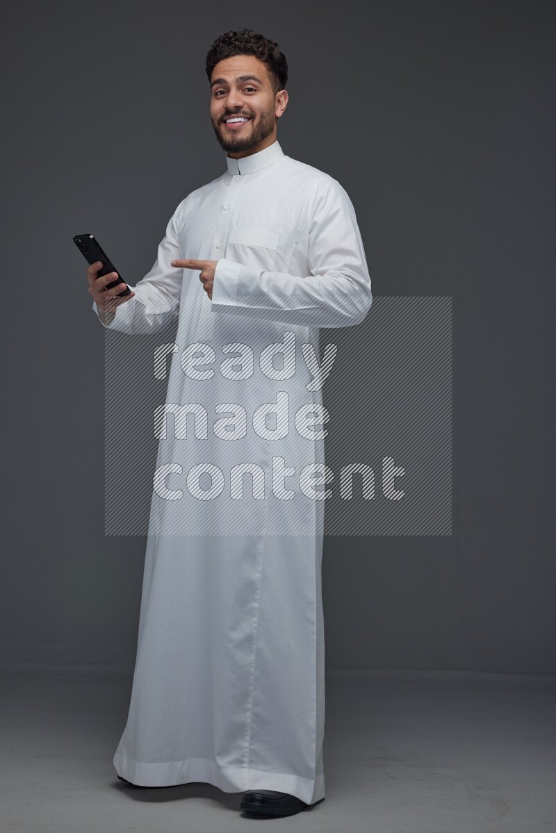 A Saudi man wearing Thobe and holding his phone while pointing to it eye level on a gray background