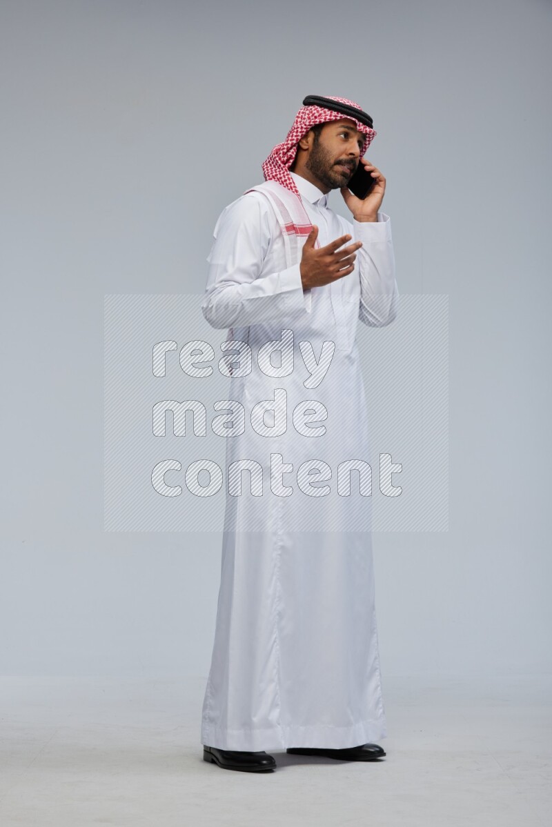 Saudi man Wearing Thob and shomag standing talking on phone on Gray background