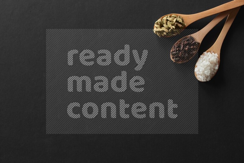 wooden spoons filled with white peppers, cloves, cardamom, salt, black peppers and basil on black flooring