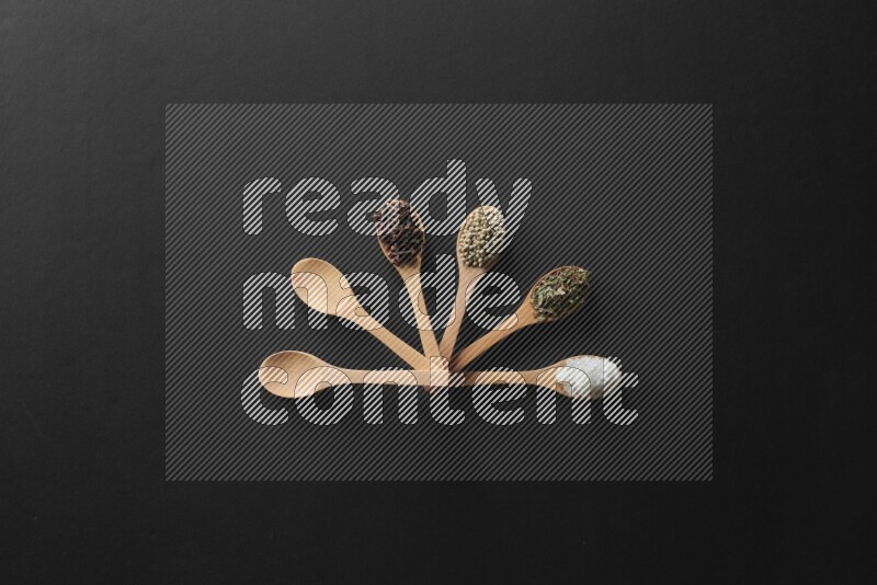 wooden spoons filled with white peppers, cloves, cardamom, salt, black peppers and basil on black flooring