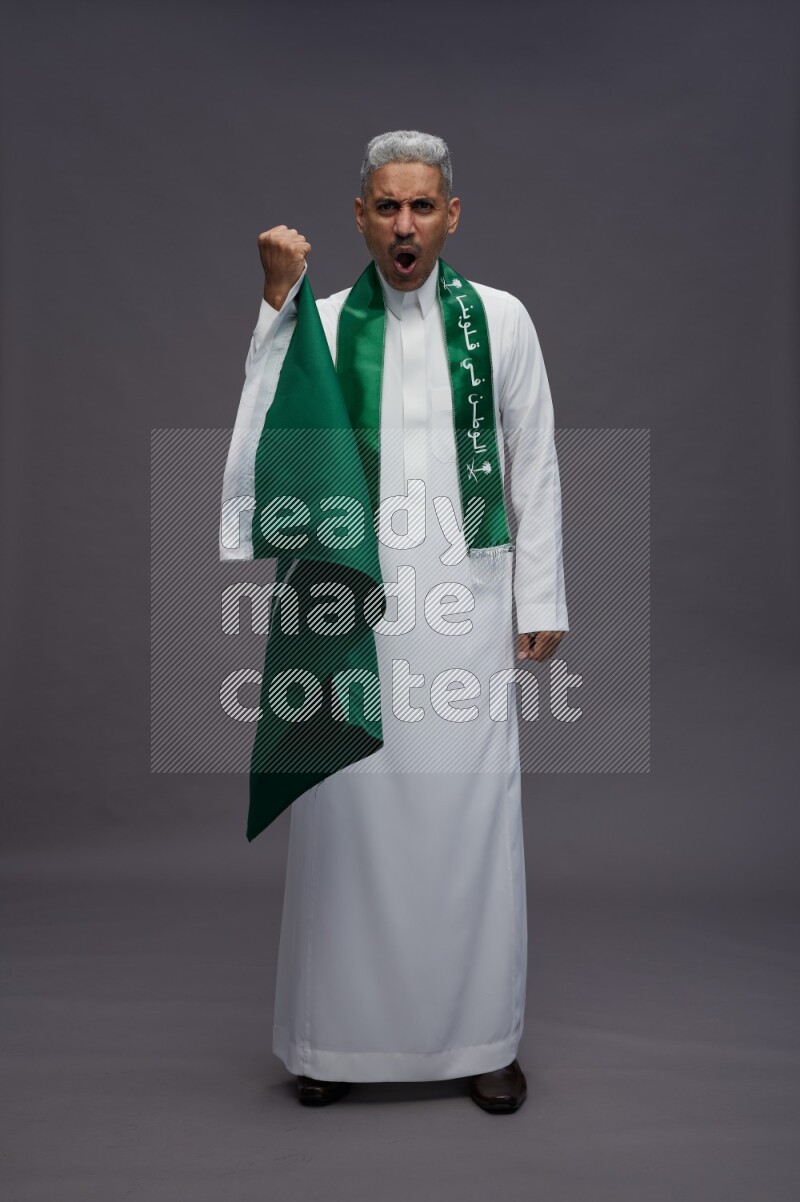 Saudi man wearing thob standing holding Saudi flag on gray background