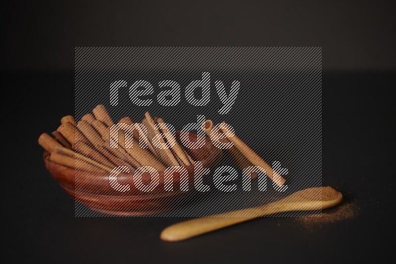 Cinnamon sticks in wooden bowl and cinnamon powder in a wooden spoon on black background
