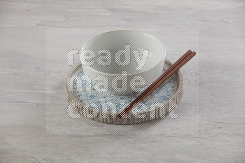 white pottery round bowl on top of multi color round ceramic plate and chopsticks, on grey textured countertop