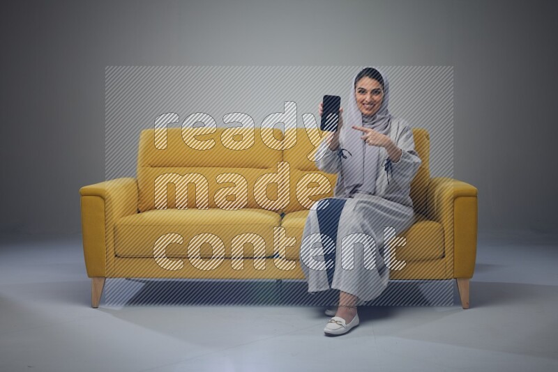 A Saudi woman wearing a light gray Abaya and head scarf setting on a yellow sofa and holding her phone while showing it's screen eye level on a grey background