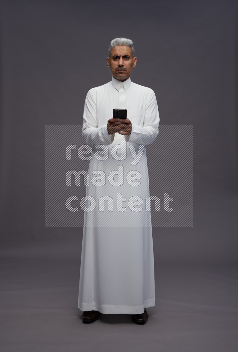 Saudi man wearing thob standing texting on phone on gray background
