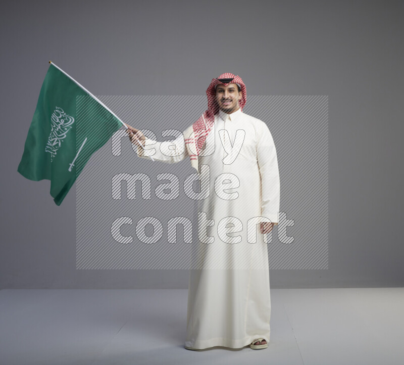 A Saudi man standing wearing thob and red shomag raising big Saudi flag on gray background