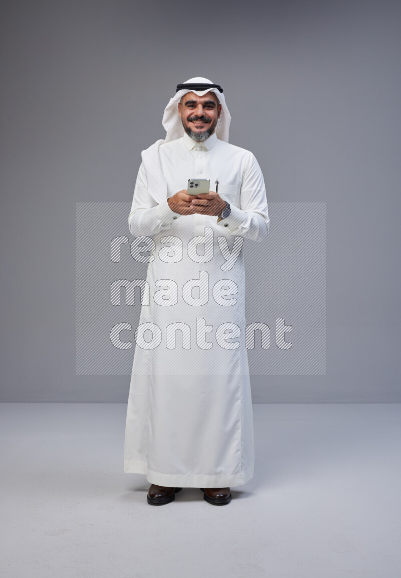 Saudi man Wearing Thob and white Shomag standing texting on phone on Gray background