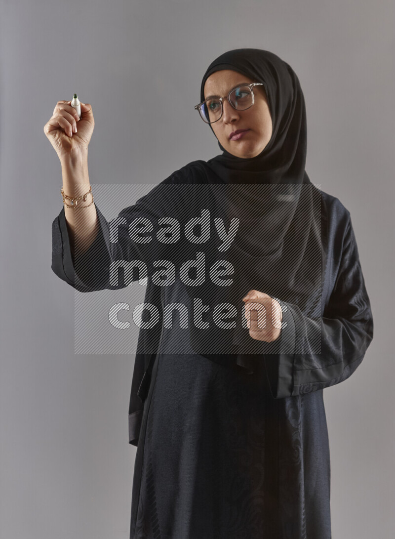 Woman in black abaya, hijab and glasses holding a marker pen to write on a transparent board with different reactions and expressions on grey background