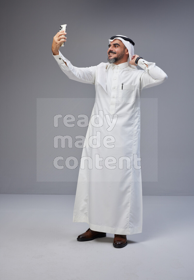 Saudi man Wearing Thob and white Shomag standing taking selfie on Gray background