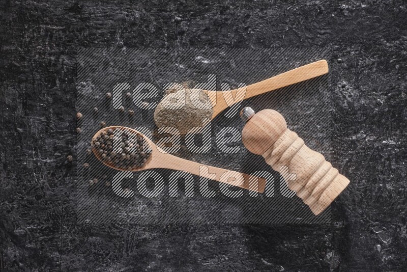 Black pepper powder and black pepper beads in wooden spoons and wooden grinder on a textured black flooring