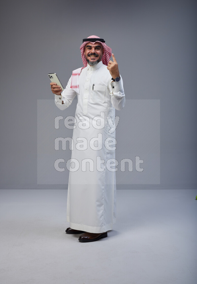 Saudi man Wearing Thob and red Shomag standing texting on phone on Gray background