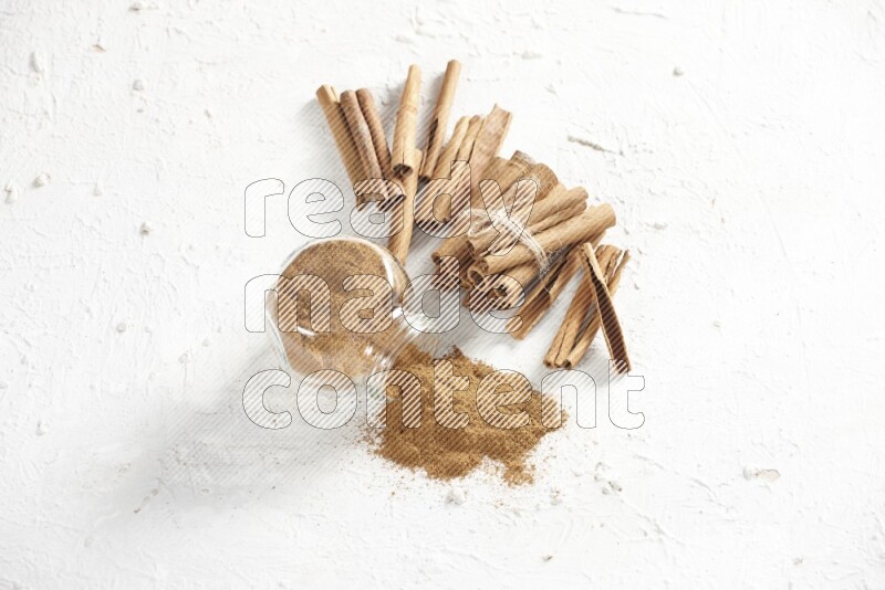 Flipped herbs glass jar full of cinnamon powder and cinnamon sticks in the back on a textured white background
