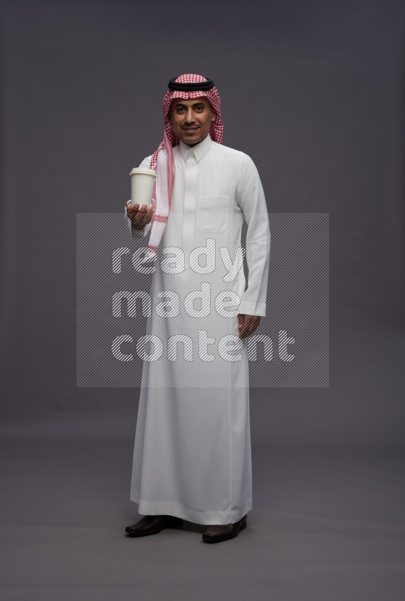 Saudi man wearing thob and shomag standing holding paper cup on gray background