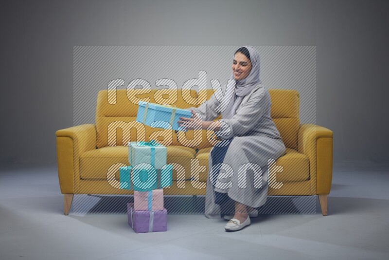 A Saudi woman wearing a light gray Abaya and head scarf sitting on a yellow sofa holding a gift box beside multi color gift boxes eye level on a grey background