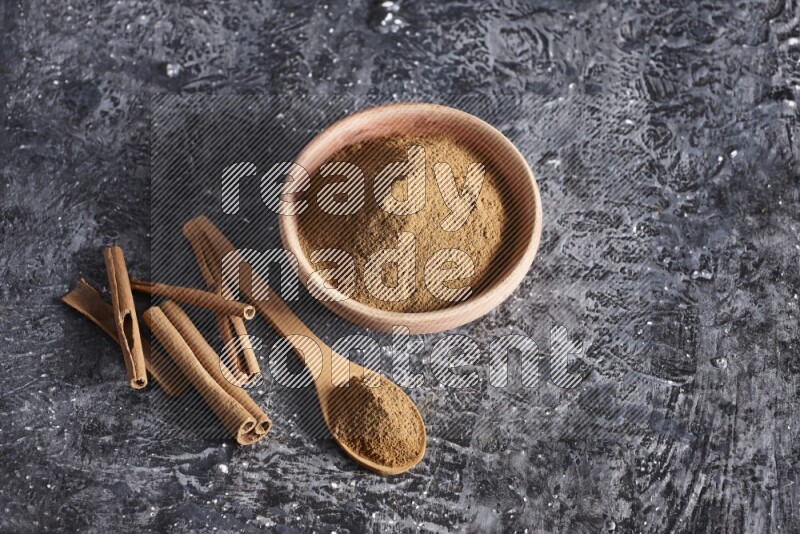 wooden bowl full of cinnamon powder and a wooden spoon full of it with cinnamon sticks on a textured black background