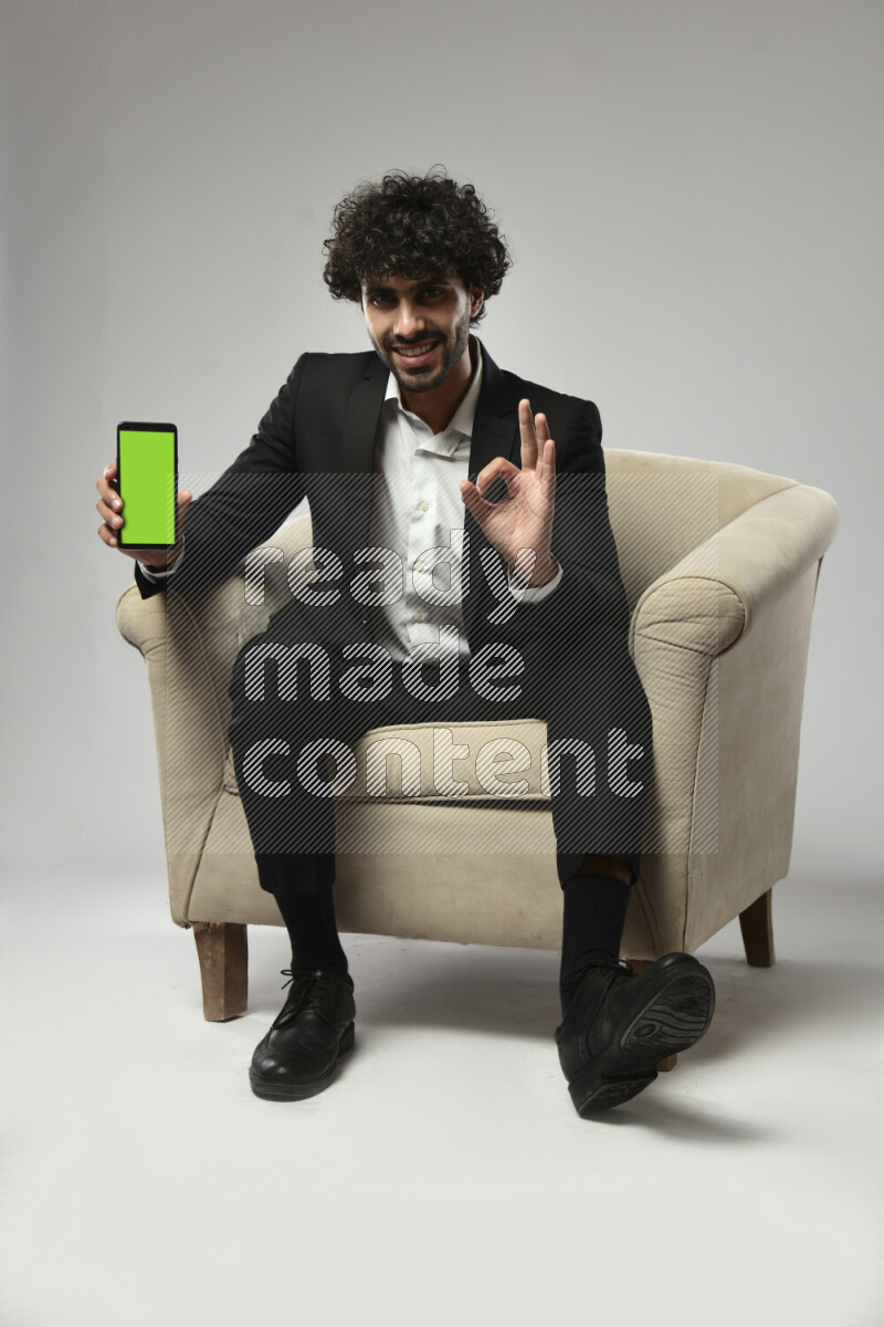 A man wearing formal sitting on a chair showing a phone screen on white background