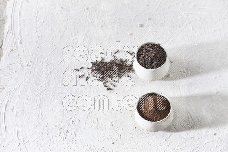 2 Beige ceramic bowls, one full of cloves and the other full of powder on textured white flooring