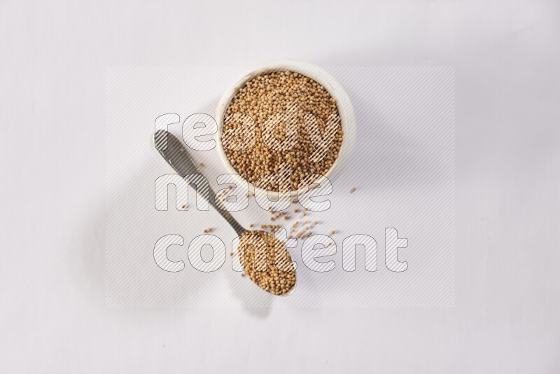A beige pottery bowl and a metal spoon full of mustard seeds on white flooring