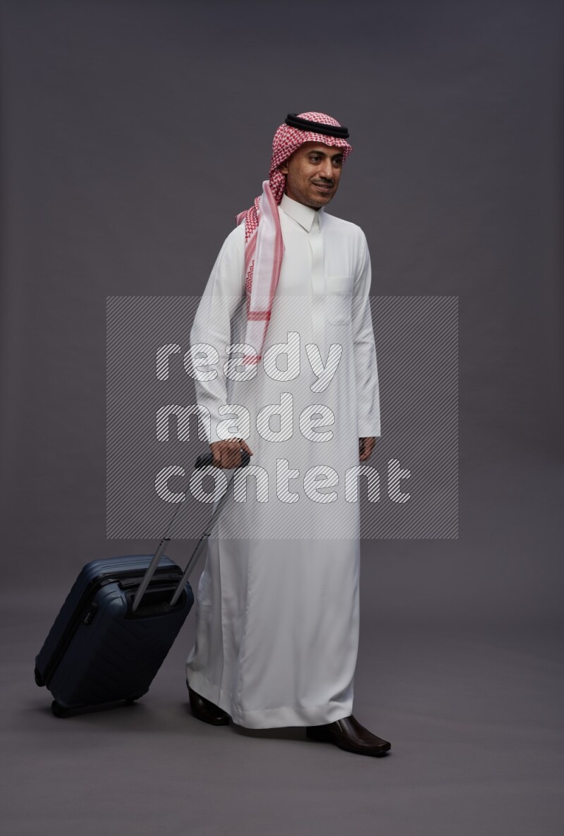 Saudi man wearing thob and shomag standing holding bag on gray background