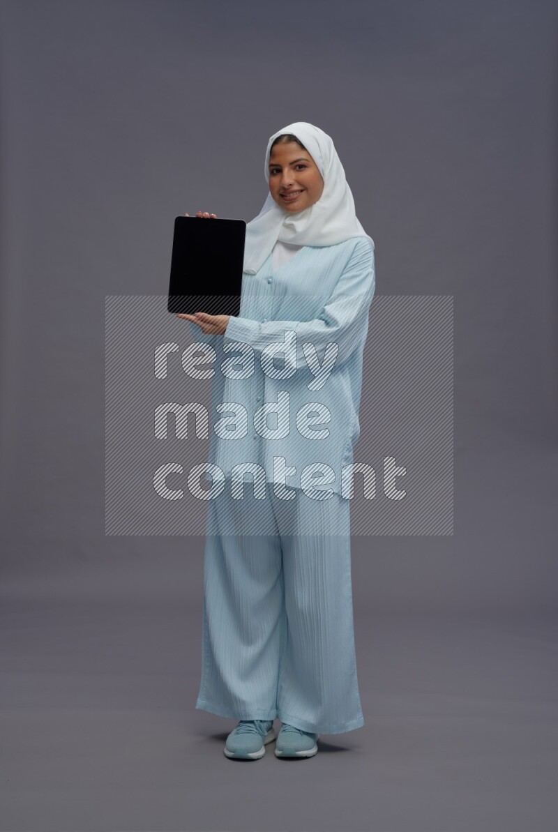 Saudi woman wearing hijab clothes standing showing tablet to camera on gray background