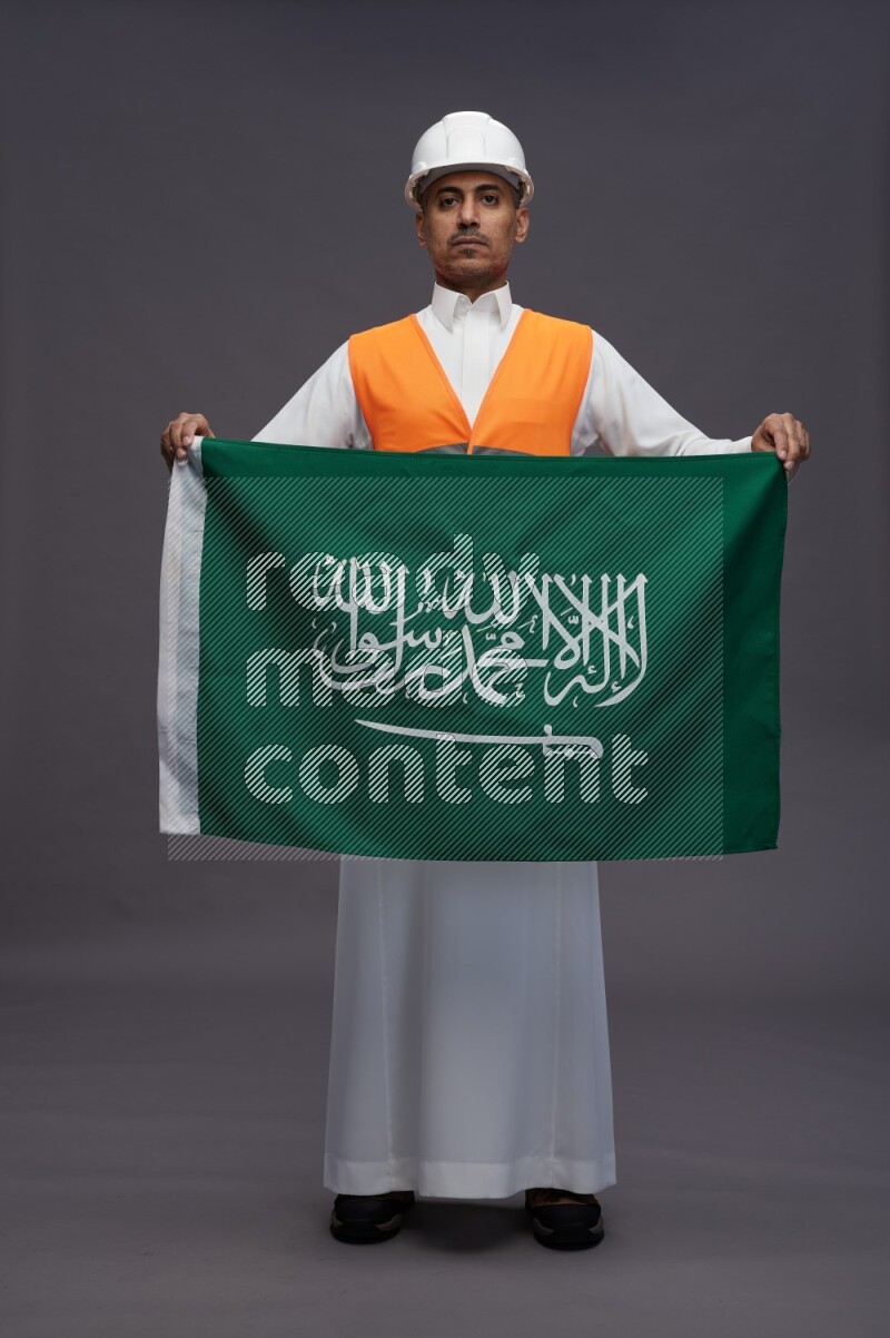 Saudi man wearing thob with engineer vest standing holding Saudi flag on gray background