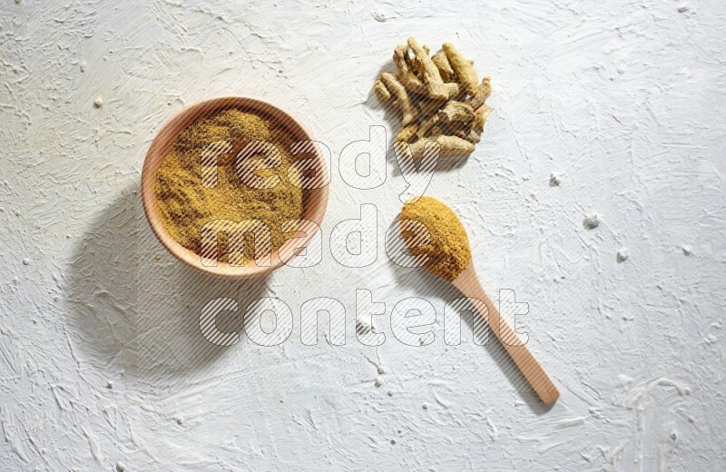 A wooden bowl and wooden spoon full of turmeric powder with dried turmeric fingers on textured white flooring