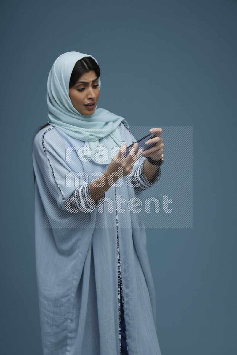 A Saudi woman Playing a Game on her phone on a blue background wearing a blue Abaya with hijab