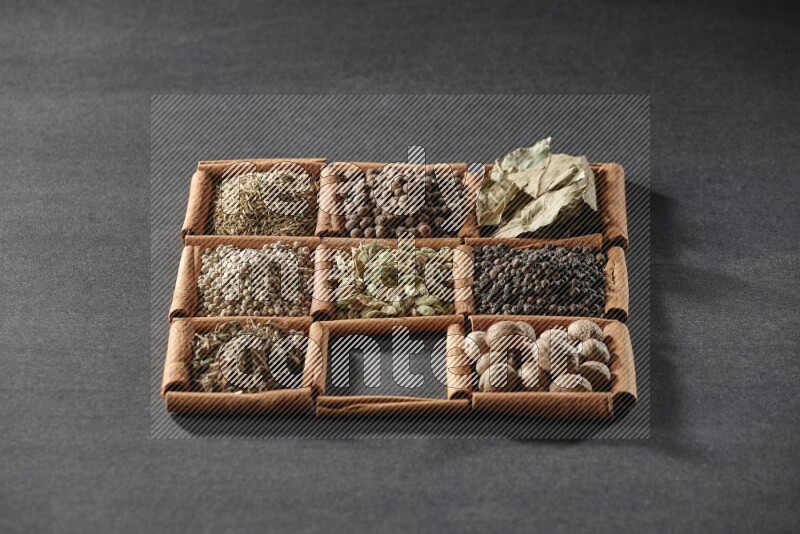 9 squares of cinnamon sticks full of cardamom in the middle surrounded by nutmeg, black pepper, bay laurel leaves, allspice, cumin, white pepper, dried basil and salt on black flooring