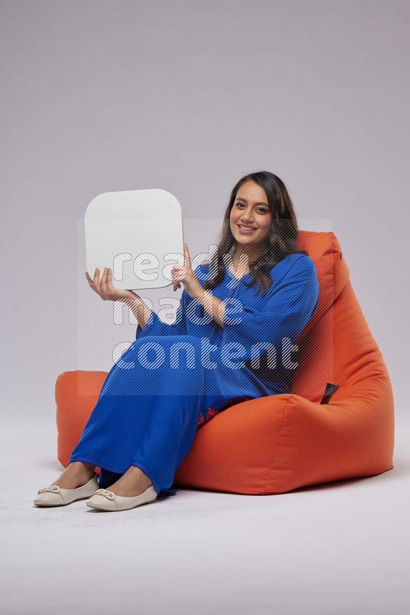 A Woman sitting on an orange beanbag wearing Jalabeya holding a social media sign