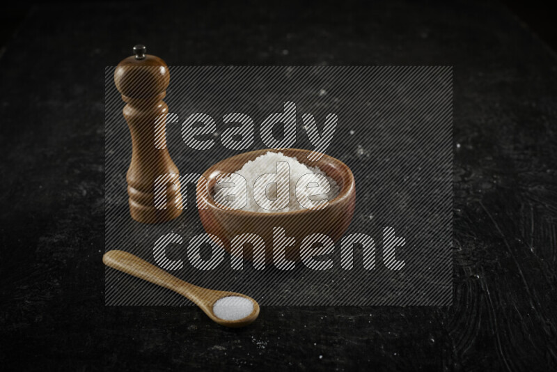A wooden bowl and spoon filled with white sea salt and wooden grinder beside them on black background
