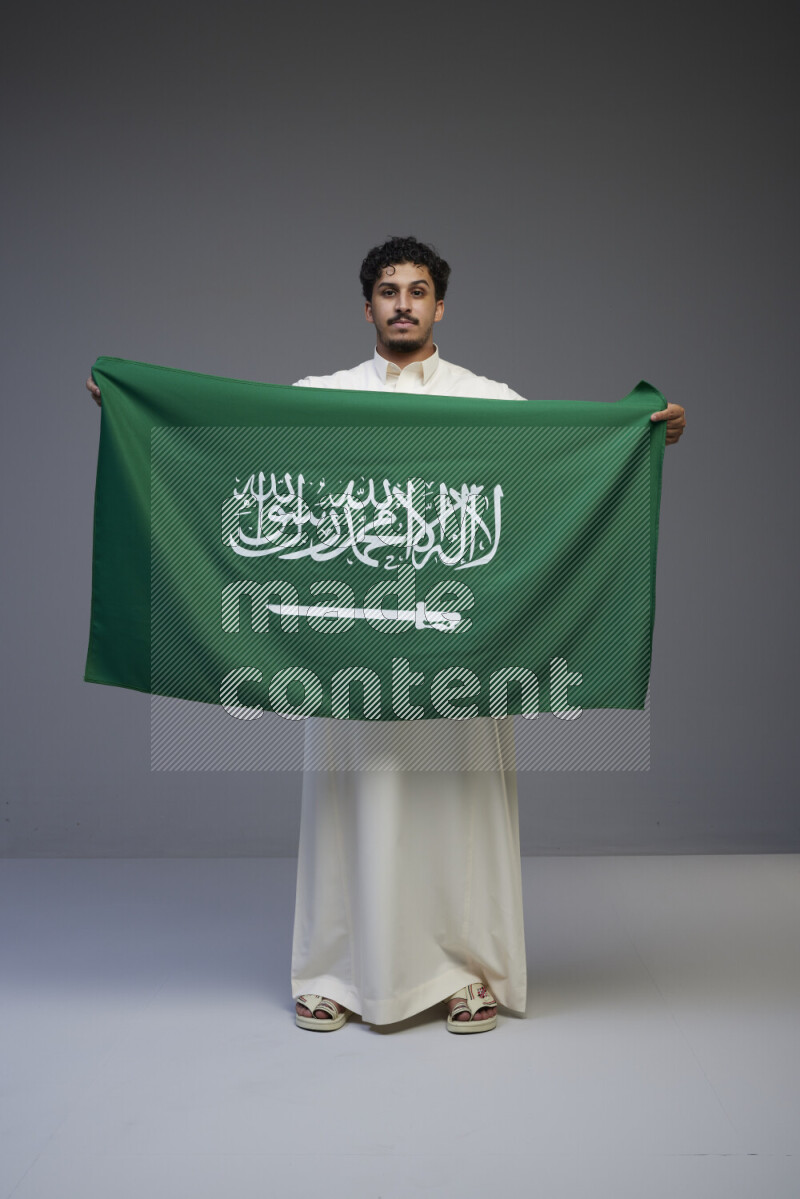A saudi man standing wearing thob holding big saudi flag on gray background