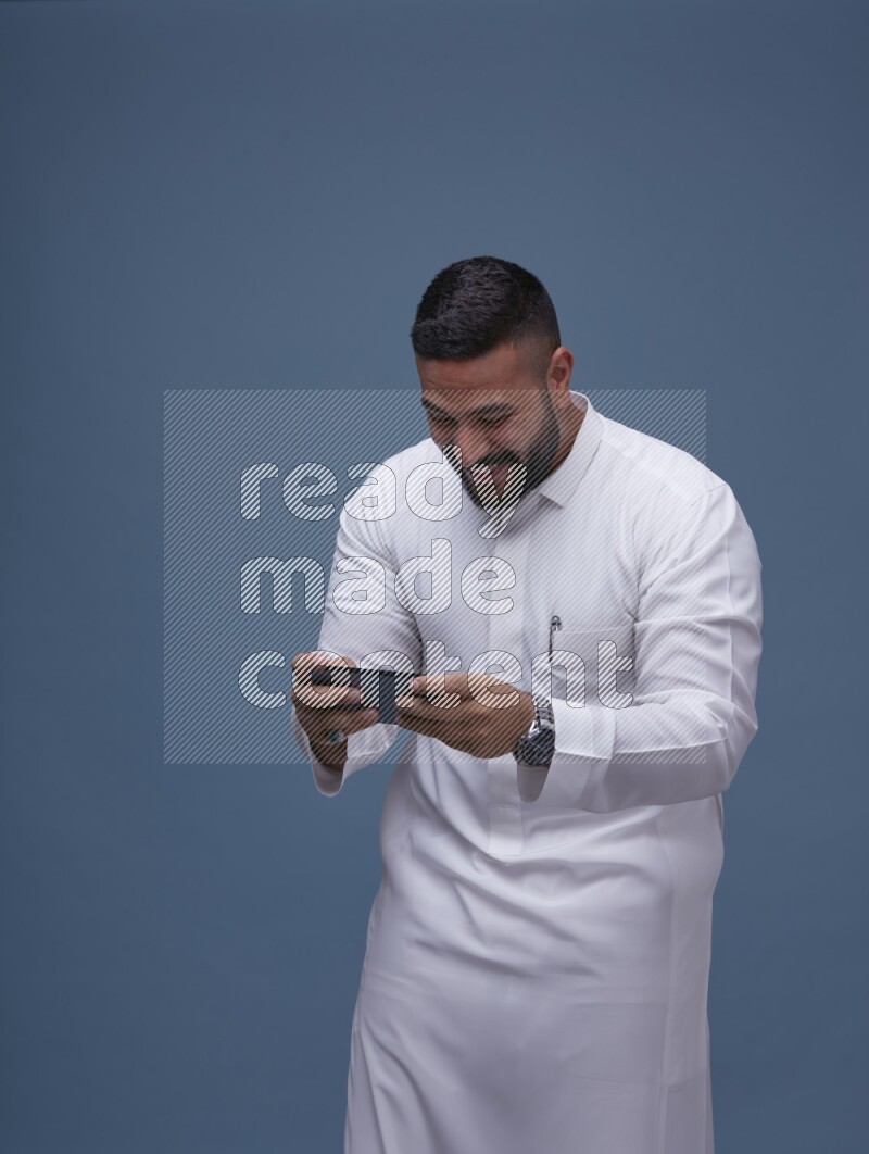 A man Playing a Game on his Smartphone  on Blue Background wearing Saudi Thob