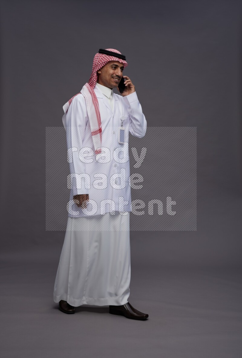 Saudi man wearing thob with lab coat and shomag with pocket employee badge standing talking on phone on gray background