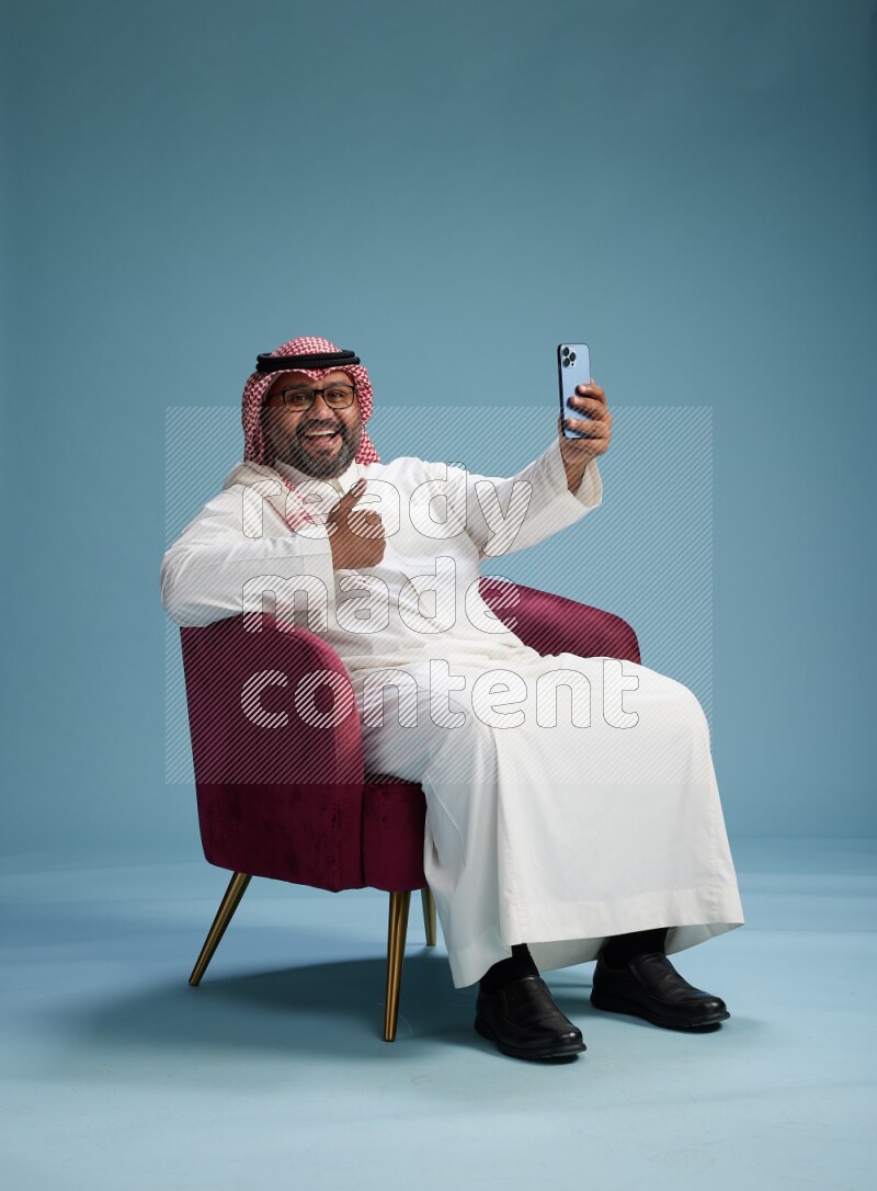 Saudi Man with shimag sitting on chair taking selfie on blue background