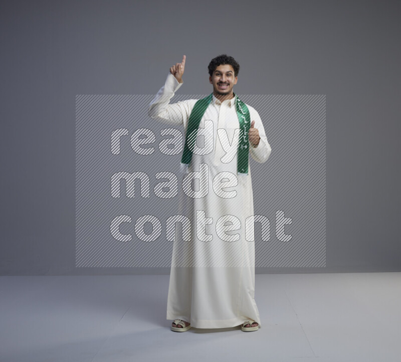 A Saudi man standing wearing thob and Saudi flag scarf on gray background