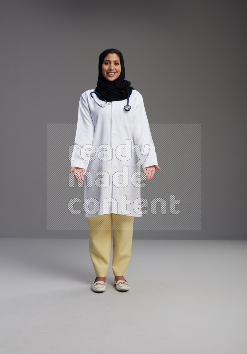 Saudi woman wearing lab coat with stethoscope standing interacting with the camera on Gray background