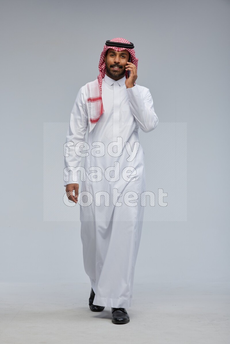 Saudi man Wearing Thob and shomag standing talking on phone on Gray background