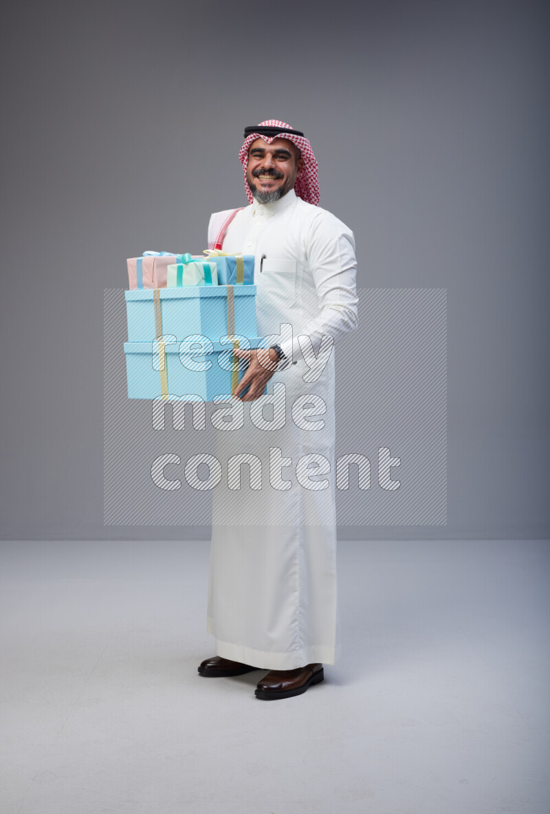 Saudi man Wearing Thob and red Shomag standing holding gift box on Gray background