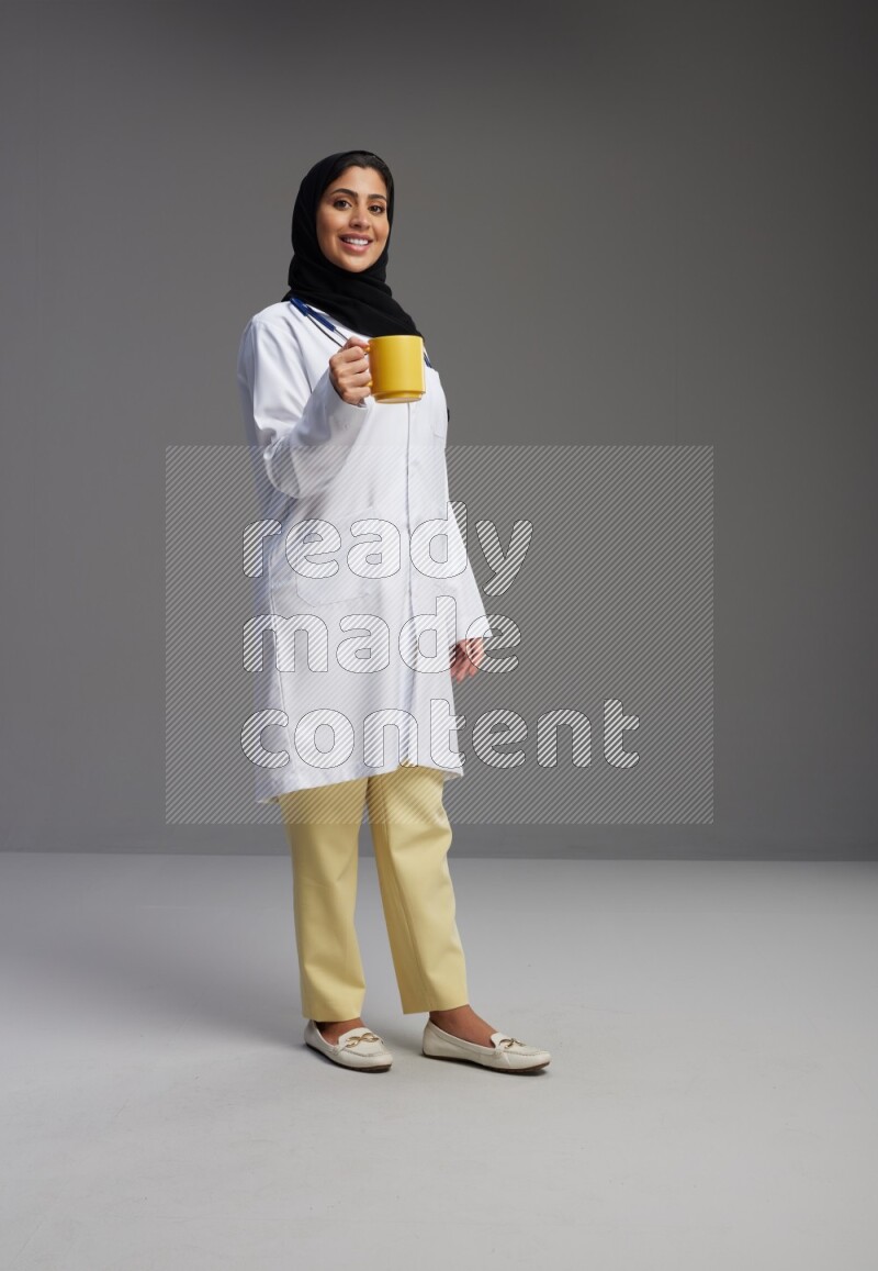 Saudi woman wearing lab coat with stethoscope standing holding mug on Gray background