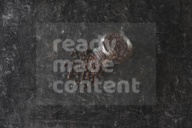 A flipped glass spice jar full of cloves and cloves came out of it on textured black flooring