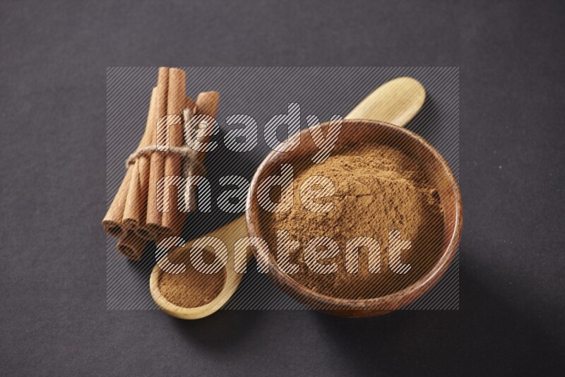 Cinnamon sticks stacked and bounded beside a wooden bowl full of cinnamon powder and a wooden spoon full of powder on black background