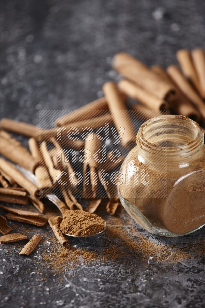 Herbal glass jar and a metal spoon full of cinnamon powder surrounded by cinnamon sticks on textured black background