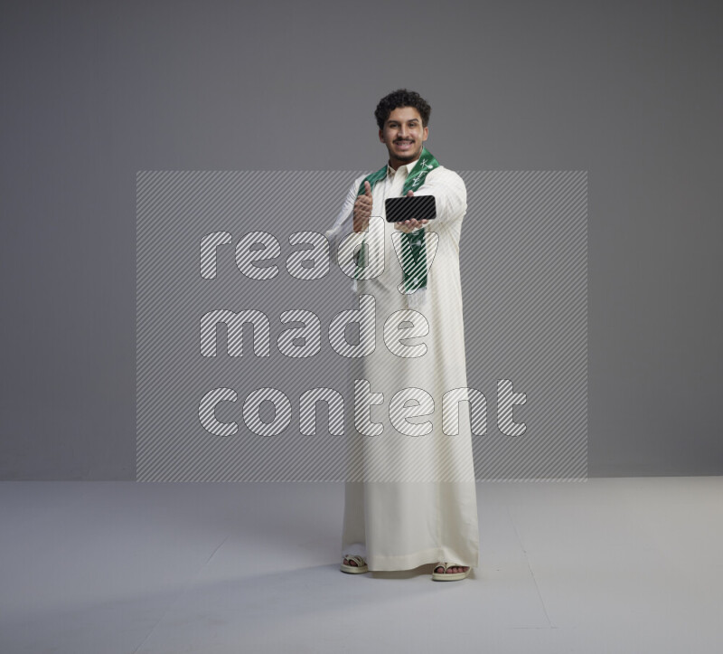 A Saudi man standing wearing thob and Saudi flag scarf showing phone to camera on gray background
