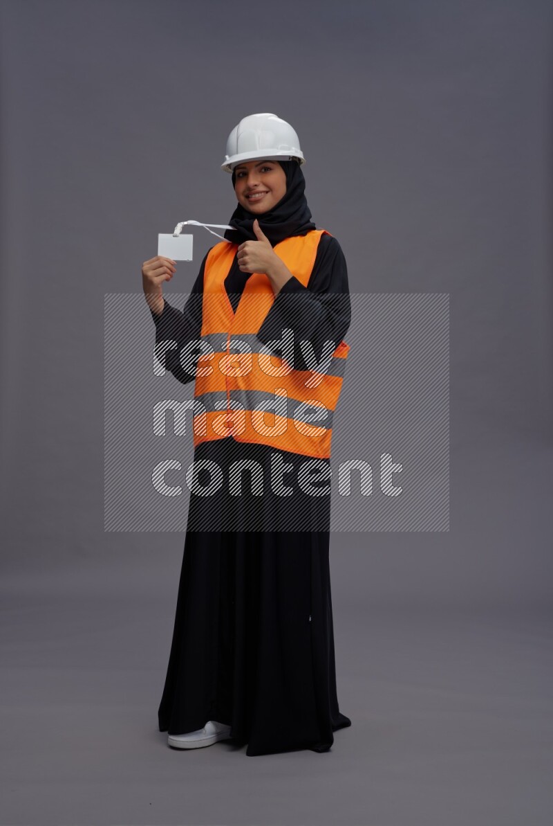 Saudi woman wearing Abaya with engineer vest with neck strap employee badge standing interacting with the camera on gray background