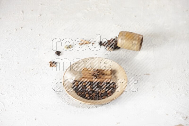 Beige plate full of coffee beans, cinnamon sticks and star anise with a coffee grinder, coffee beans, cinnamon pieces and cardamom next of it on white background