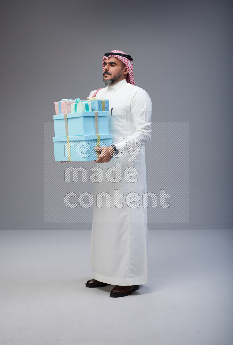 Saudi man Wearing Thob and red Shomag standing holding gift box on Gray background